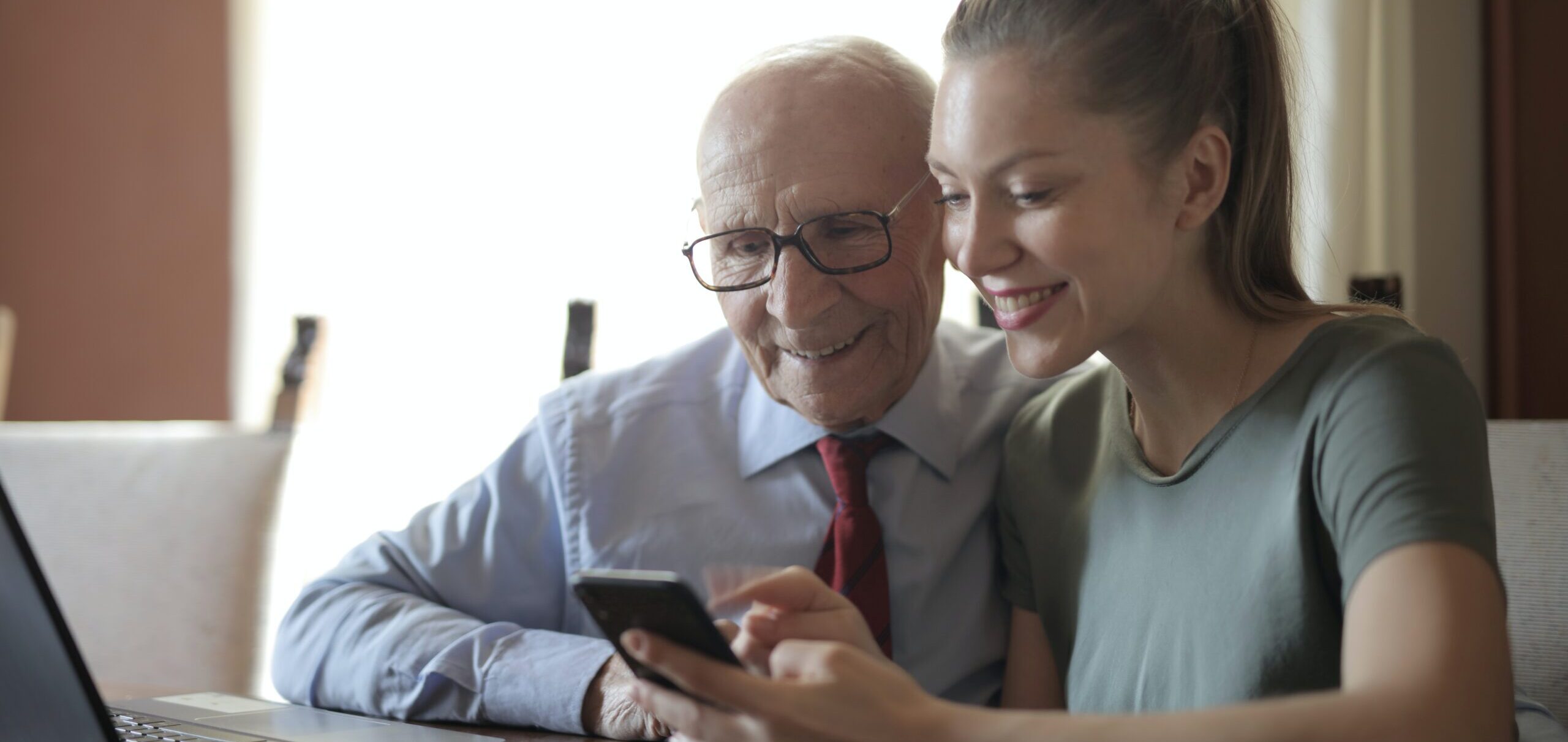An older person sits with a younger person and they look at a smartphone together.
