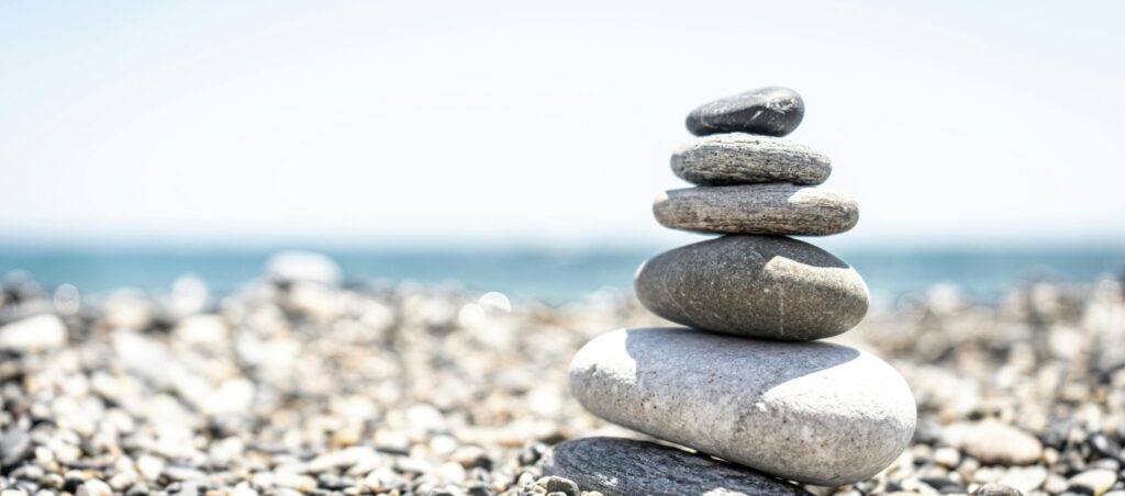 A balanced stack of stones sit on a pebble beach on front of water in the sun