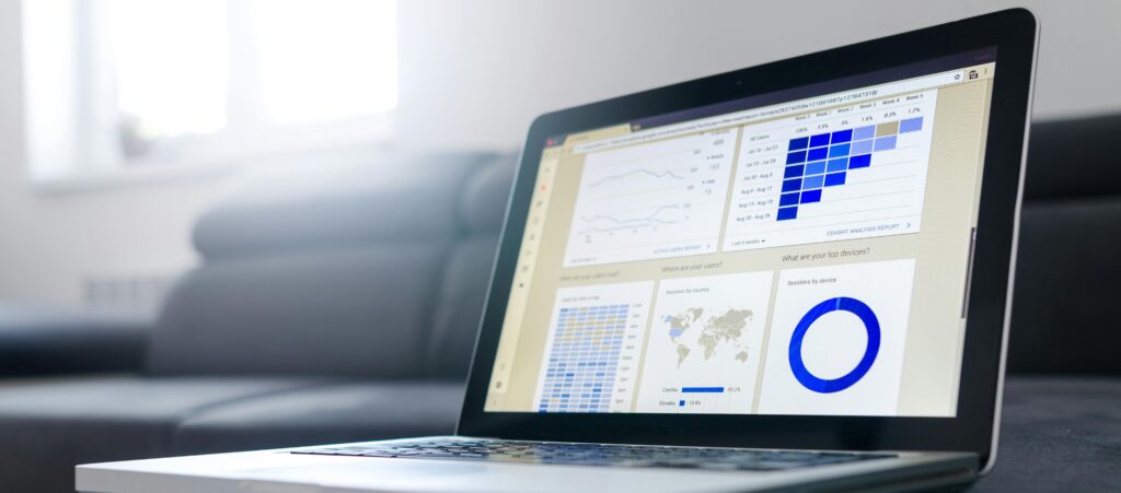 An illuminated laptop sits on a grey desk. The screen shows a Google Analytics 4 report.